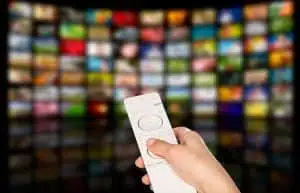 Hand holding remote control with blurred TV screens background.